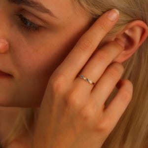 May include: A gold band ring with five small, colorful gemstones. The gemstones are set in a row and are a mix of blue, red, and white.