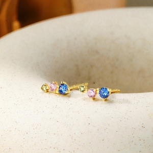 Puede incluir: Un par de pendientes de botón de oro con un grupo de piedras preciosas azules, rosas y verdes. Los pendientes son pequeños y delicados, perfectos para el uso diario.