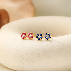 May include: A pair of gold stud earrings with a flower design. The earrings have a center stone surrounded by smaller stones. One earring has blue stones and the other has red stones.