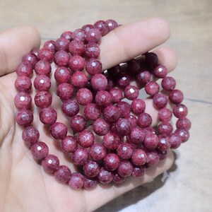 May include: A strand of faceted, deep magenta-coloured gemstone beads. The beads are round, polished, and feature a multi-faceted surface that reflects light. The strand is coiled into a circular shape, showcasing the consistent dark pink hue.