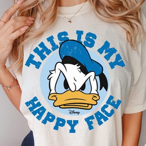 May include: Ivory-colored t-shirt featuring a graphic of Donald Duck with the text "THIS IS MY HAPPY FACE" in blue. The design includes Donald Duck's face inside a light blue circle. The Disney logo is at the bottom.