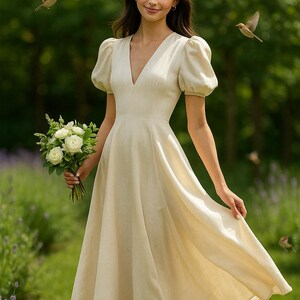 May include: A woman in a cream-colored linen dress with a v-neck and puffed sleeves. She is holding a bouquet of white flowers and is standing in a garden.