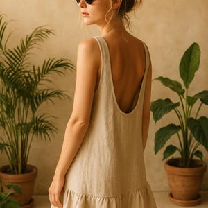 May include: A beige sleeveless dress with a low back and a ruffled hem. The person in the image is wearing black sunglasses. Potted plants are in the background.