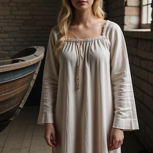 K&ouml;nnte beinhalten: Ein wei&szlig;es, lang&auml;rmeliges Baumwollkleid mit quadratischem Ausschnitt und einem Kordelzug am Hals. Das Kleid hat ein schlichtes, elegantes Design.