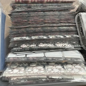 May include: A clear plastic storage container filled with stacks of various patterned and plain adhesive sheets. The sheets are organized by color and design, and include hearts, flowers, and other decorative elements.