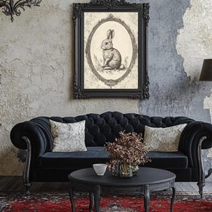 May include: A black velvet tufted sofa with two white throw pillows sits in front of a gray wall with a black framed print of a rabbit. A black lamp with a black shade stands to the right of the sofa. A black coffee table with a small white bowl and a vase of dried flowers sits in front of the sofa.