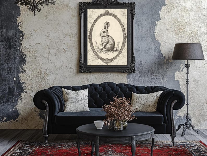 May include: A black velvet tufted sofa with two white throw pillows sits in front of a gray wall with a black framed print of a rabbit. A black lamp with a black shade stands to the right of the sofa. A black coffee table with a small white bowl and a vase of dried flowers sits in front of the sofa.