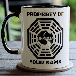 May include: White ceramic mug with a black handle and rim. The mug features a black Dharma Initiative logo with the text "PROPERTY OF" and "YOUR NAME".