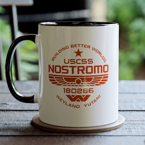 May include: White ceramic mug with a black handle and rim, featuring the text "USCSS NOSTROMO" in orange, along with other text and a graphic. The mug sits on a coaster on a wooden table.