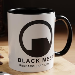 May include: White ceramic mug with a black rim and handle. The mug features a black and white logo of a circle with a rectangle inside, and the text "BLACK MESA RESEARCH FACILITY".