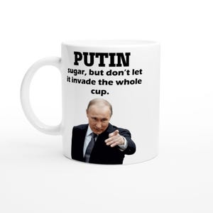 Op de afbeelding: Witte keramische mok met het woord "PUTIN" in vette zwarte letters. De mok toont ook de zin "sugar, but don't let it invade the whole cup." Een afbeelding van een man in een pak staat eronder.