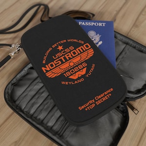 Op de afbeelding: Zwarte reiszak met oranje tekst en afbeeldingen. De portemonnee heeft de tekst "USCSS NOSTROMO" en "Security Clearance *TOP SECRET*". Een blauw paspoort is zichtbaar in de open portemonnee. De portemonnee heeft een schouderriem.