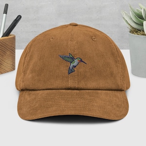 Puede incluir: Una gorra de béisbol de pana marrón con un diseño de colibrí bordado. El colibrí es verde, azul y rojo.