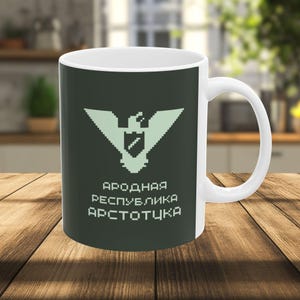 May include: White ceramic mug with a dark green background and a pixelated eagle emblem. The mug features text in a foreign language. The mug is sitting on a wooden surface.
