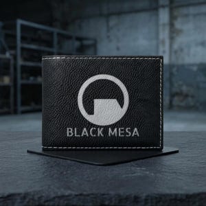 Może przedstawiać: Czarny portfel z białymi przeszyciami i napisem "BLACK MESA" pod białym logo. Portfel wykonany jest z czarnego, teksturowanego materiału i leży na czarnym kwadracie. Tło to rozmyte otoczenie przemysłowe.
