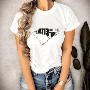 May include: White t-shirt with a black outline of the state of North Carolina and the text "It's Better On Top" printed on it.