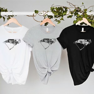 May include: Three t-shirts with a graphic of the state of North Carolina and the text "It's Better On Top". The shirts are white, grey, and black.