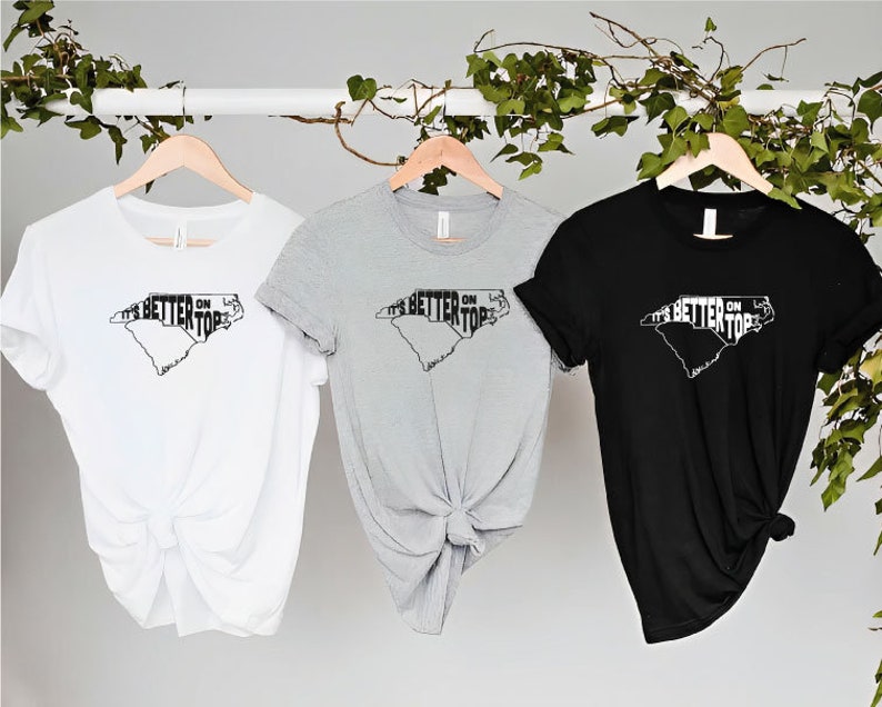 May include: Three t-shirts with a graphic of the state of North Carolina and the text "It's Better On Top". The shirts are white, grey, and black.