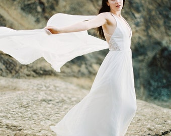 May include: A woman in a white flowing dress with a sheer overlay stands on a rocky beach. The dress has a V-neckline and a lace detail at the bodice. The woman is looking at the camera with a slight smile.