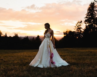 May include: A woman in a white wedding dress with colorful floral accents stands in a field at sunset. The dress has a strapless bodice and a flowing skirt.