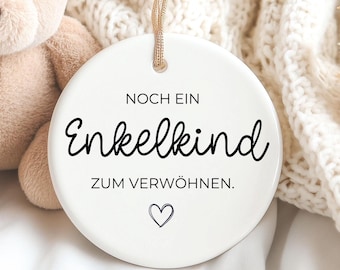 Pode incluir: Um urso de pelúcia bege está ao lado de um enfeite branco e redondo. O enfeite tem o texto "NOCH EIN Enkelkind ZUM VERWÖHNEN." e um símbolo de coração. O enfeite está pendurado por uma corda bege. O fundo é um cobertor branco.