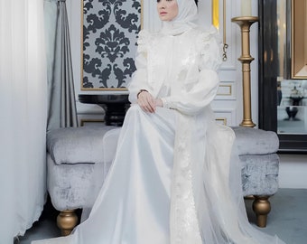 May include: A white long-sleeved dress featuring a satin skirt and a sheer overlay with ruffled shoulders. The garment is paired with a matching white headscarf. The dress has a structured bodice and a flowing floor-length hemline with subtle detailing.