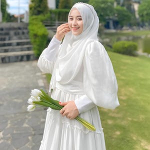 Puede incluir: Una mujer con un vestido blanco y un hiyab blanco con una diadema de perlas. Lleva un ramo de tulipes blancas.
