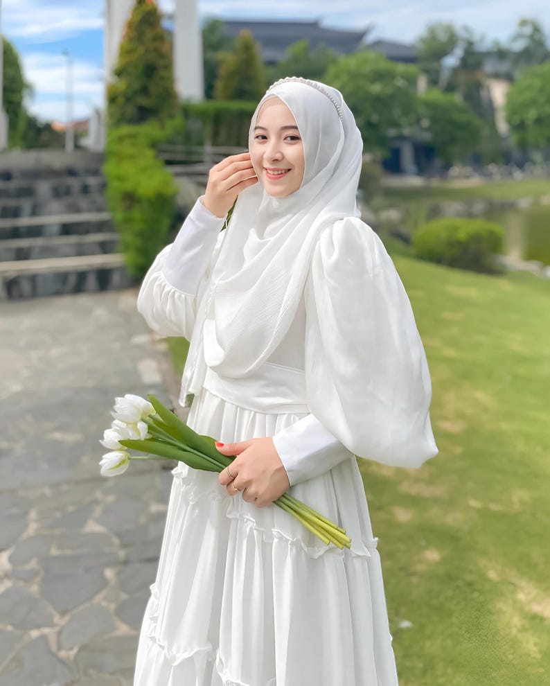 Puede incluir: Una mujer con un vestido blanco y un hiyab blanco con una diadema de perlas. Lleva un ramo de tulipes blancas.