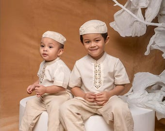 Puede incluir: Dos niños pequeños con conjuntos crema a juego, que incluyen camisas bordadas, pantalones y sombreros. Están sentados en una plataforma blanca, con sandalias marrones. El fondo es de color marrón cálido. Adecuado para una ocasión especial.