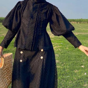 Puede incluir: Un conjunto de top y falda de encaje negro con botones dorados. El top tiene cuello alto y mangas largas con hombros abullonados. La falda es de línea A y llega a las rodillas.