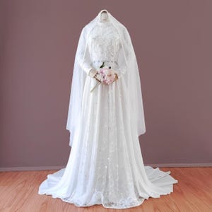 Peut inclure: Une robe de mariée blanche à manches longues et un voile. La robe a un corsage ajusté avec des détails en dentelle et une jupe ample. La mariée tient un bouquet de fleurs roses.