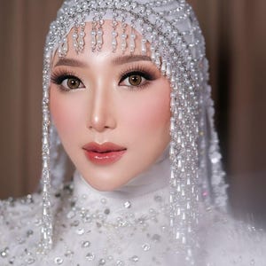 Op de afbeelding: Een vrouw die een witte bruidsjurk draagt met een parelversierd hoofdtooi. De hoofdtooi heeft een lange, parelversierde franje die langs de achterkant van haar nek hangt. De jurk is bedekt met kleine, witte pailletten.