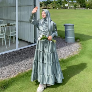 Puede incluir: Una mujer con un vestido maxi verde claro con falda de volantes y un pa&ntilde;uelo a juego. Est&aacute; de pie en una zona de c&eacute;sped con un edificio blanco al fondo.