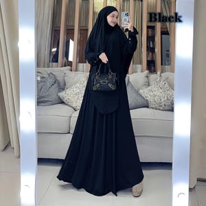 Può includere: Un'abaya nera con una gonna lunga e fluida e un velo abbinato. L'abaya ha una piccola borsa nera con dettagli dorati.
