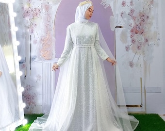 May include: A floor-length white gown featuring long sleeves, a high neckline, and an all-over sequin embellishment. The dress includes a pearl-accented waist belt and a sheer tulle cape overlay. A matching white hijab and a gold crown are worn.