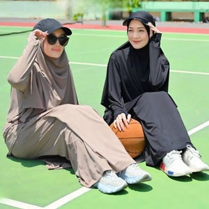 Può includere: Due persone con abbigliamento sportivo modesto, tra cui berretti, occhiali da sole e top e gonne a maniche lunghe, sono sedute su una superficie verde. Una è color talpa, l'altra nera. Un pallone da basket è tra loro. Entrambe indossano scarpe sportive.