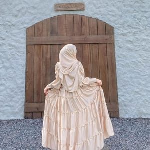 Puede incluir: Una mujer lleva un vestido largo de color crema con volantes y un pa&ntilde;uelo a juego. El vestido tiene un dobladillo con volantes y la mujer est&aacute; de pie frente a una puerta de madera con un letrero que dice "Barn Yard".