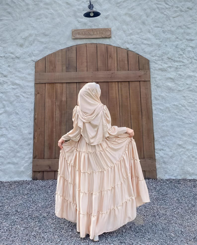 Puede incluir: Una mujer lleva un vestido largo de color crema con volantes y un pa&ntilde;uelo a juego. El vestido tiene un dobladillo con volantes y la mujer est&aacute; de pie frente a una puerta de madera con un letrero que dice "Barn Yard".