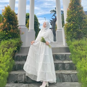 Puede incluir: Una mujer con un hiyab blanco y un vestido blanco con falda de volantes. Lleva un ramo de flores verdes y est&aacute; de pie en un conjunto de escalones de piedra.