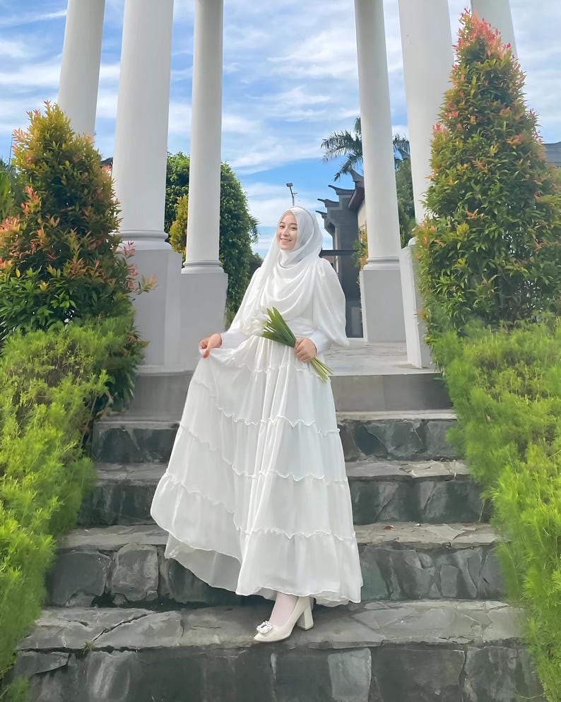 Puede incluir: Una mujer con un hiyab blanco y un vestido blanco con falda de volantes. Lleva un ramo de flores verdes y est&aacute; de pie en un conjunto de escalones de piedra.