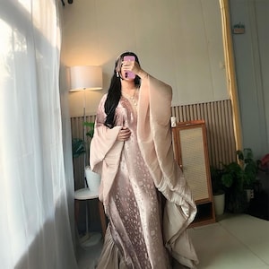 以下が含まれることがあります： モーヴとシャンパンの柔らかな色合いのフルレングスのカフタンドレス。サテンのような生地に繊細な同系色の花柄が施され、幅広で流れるような袖と、ネックラインには複雑なゴールドトーンの刺繍があしらわれています。