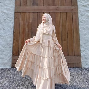 Puede incluir: Una mujer lleva un vestido largo de color beige con volantes y un pa&ntilde;uelo a juego. El vestido tiene un corpi&ntilde;o ajustado y una falda fluida con varias capas. Est&aacute; de pie frente a una puerta de madera.