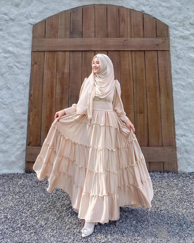 Puede incluir: Una mujer lleva un vestido largo de color beige con volantes y un pa&ntilde;uelo a juego. El vestido tiene un corpi&ntilde;o ajustado y una falda fluida con varias capas. Est&aacute; de pie frente a una puerta de madera.