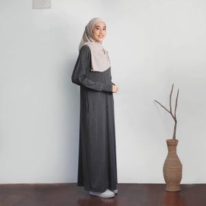 May include: A woman wearing a long-sleeved, charcoal gray maxi dress with a light beige headscarf. She is standing in front of a white wall with a vase and a branch in the background.