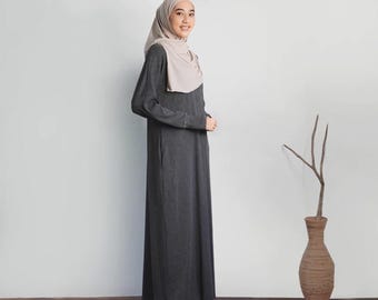 Puede incluir: Una mujer con un vestido largo de manga larga de color gris antracita y un pañuelo de cabeza beige claro. Está de pie frente a una pared blanca con un jarrón y una rama en el fondo.