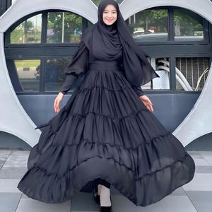 Puede incluir: Un vestido negro maxi con falda de capas y mangas largas. El vestido tiene un corpi&ntilde;o ajustado y una falda fluida. La modelo lleva un pa&ntilde;uelo negro en la cabeza.
