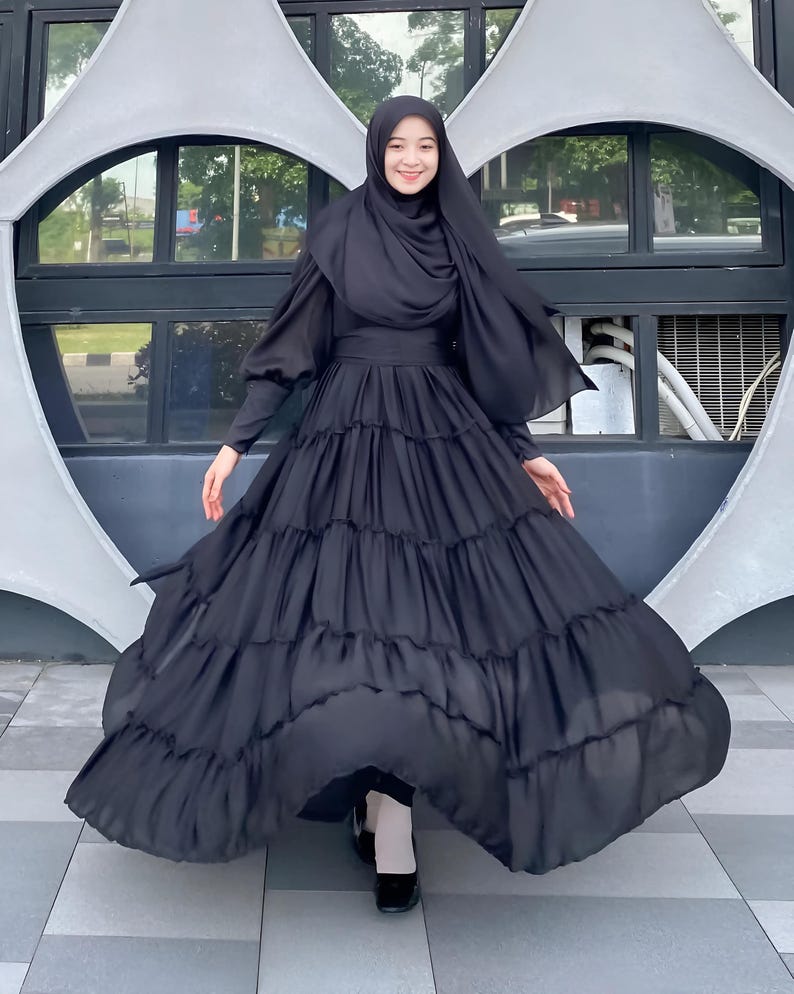 Puede incluir: Un vestido negro maxi con falda de capas y mangas largas. El vestido tiene un corpi&ntilde;o ajustado y una falda fluida. La modelo lleva un pa&ntilde;uelo negro en la cabeza.