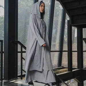 Puede incluir: Una abaya con capucha gris claro y falda a juego. La abaya tiene una parte delantera abotonada y mangas largas. La persona lleva un velo gris y zapatillas deportivas blancas y negras. El conjunto está hecho de una tela suave y fluida.