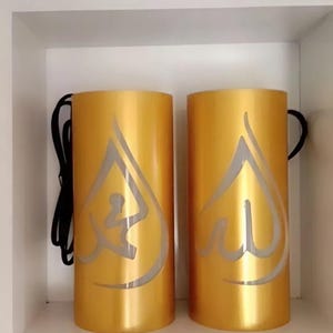 Könnte beinhalten: Zwei goldfarbene zylindrische Lampen mit einem silbernen arabischen Kalligraphie-Design. Jede Lampe hat einen schwarzen Griff und ein Kabel. Die Lampen werden auf einem weißen Regal präsentiert.