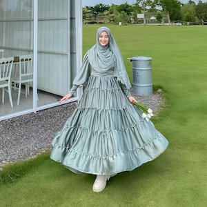 Puede incluir: Una mujer con un vestido maxi verde salvia con falda de volantes y un pa&ntilde;uelo a juego. Est&aacute; de pie en una zona de c&eacute;sped con un edificio blanco al fondo.